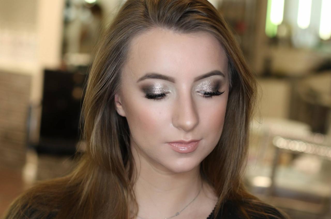 Portret kobiety z profesjonalnym makijażem oczu w odcieniach srebra i brązu, delikatny błyszczyk na ustach, włosy w odcieniach blondu i brązu, subtelna biżuteria na szyi, rozmyte tło salonu.