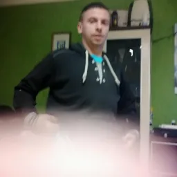 Mężczyzna w bluzie z kapturem na tle zielonej ściany, z rozmytym pierwszym planem. Widoczna szafka i obraz na ścianie.