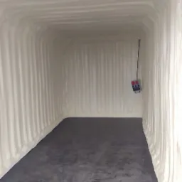 Wnętrze kontenera wykończone grubą warstwą białej pianki izolacyjnej na ścianach i suficie, z widoczną szarą podłogą i wystającym kablem z boku.