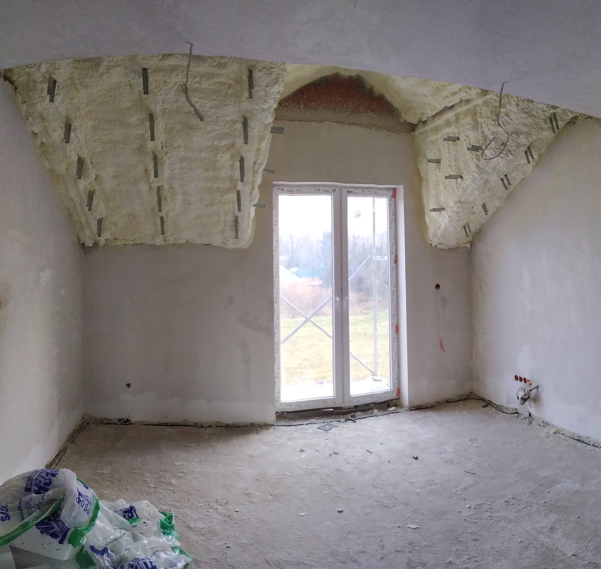 Poddasze w trakcie izolacji pianką, widoczne metalowe mocowania, okno balkonowe z widokiem na drzewa, worki z materiałem izolacyjnym na podłodze.