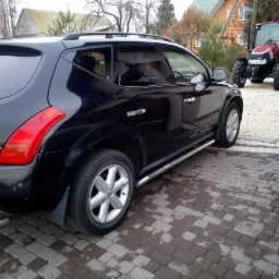 Czarny SUV Nissan Murano zaparkowany na brukowanym podjeździe, w tle czerwony traktor.