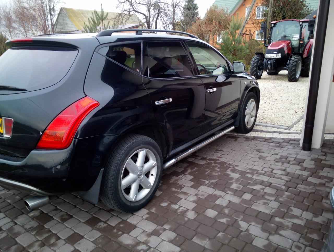 Czarny SUV Nissan Murano zaparkowany na brukowanym podjeździe, w tle czerwony traktor.