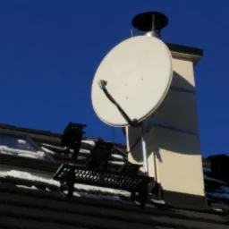 Biała antena satelitarna zamontowana na kominie domu z ciemną dachówką, widoczne fragmenty czarnego uchwytu anteny i resztki śniegu na dachu w słoneczny dzień.