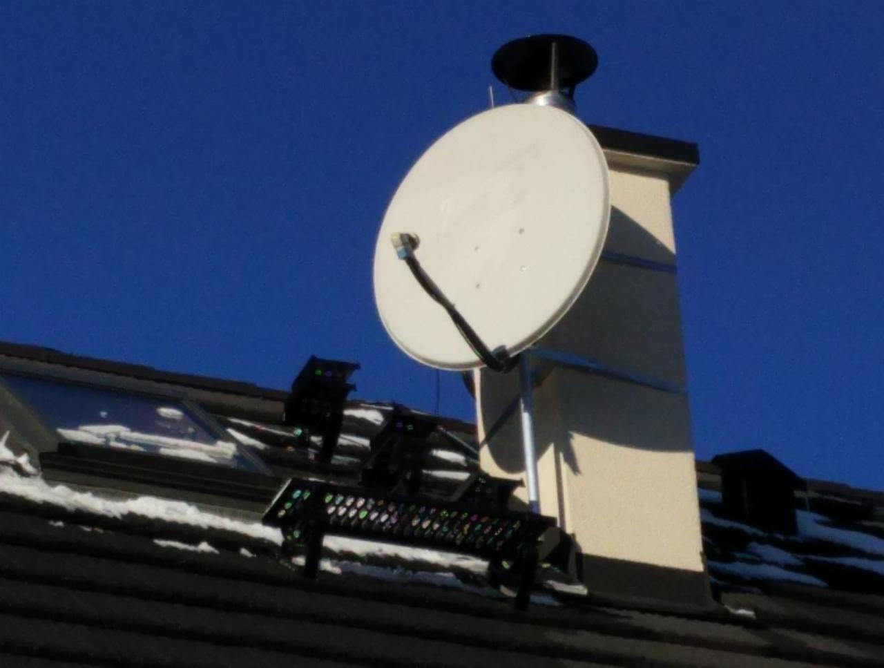 Biała antena satelitarna zamontowana na kominie domu z ciemną dachówką, widoczne fragmenty czarnego uchwytu anteny i resztki śniegu na dachu w słoneczny dzień.