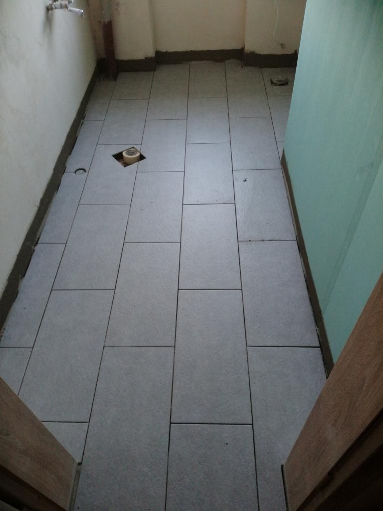 Ułożone szare płytki podłogowe w małej łazience, widoczny otwór na odpływ zabezpieczony taśmą malarską.