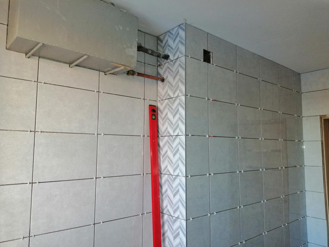 Ściana w trakcie wykańczania płytkami w odcieniach szarości, z dekoracyjnym pasem w zygzaki, widoczny poziomica i plastikowe krzyżyki dystansowe.
