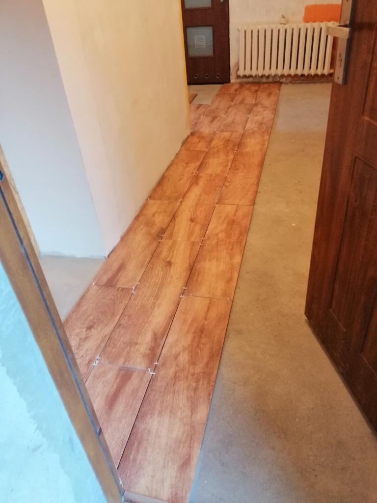 Układanie podłogi z płytek imitujących drewno w korytarzu, widoczne krzyżyki dystansowe, surowy beton na podłodze obok ułożonych płytek i grzejnik żeberkowy w tle.