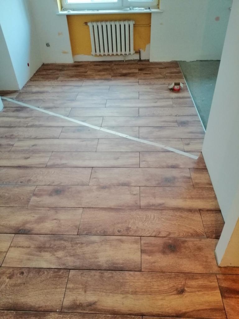 Układanie podłogi z płytek imitujących drewno, widoczna taśma maskująca i fragment nieukończonej podłogi.