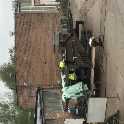 Pracownicy w odblaskowych kamizelkach pracują nad metalową konstrukcją na zewnątrz budynku z cegły, widoczne elementy spawania.