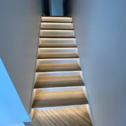 Nowoczesne schody drewniane z podświetleniem LED wzdłuż stopni, widok z dołu na górę, białe ściany, parkiet na podłodze.