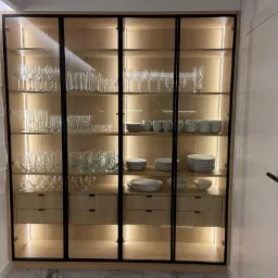 Wysoka, przeszklona szafa z drewnianymi szufladami i wewnętrznym oświetleniem, wypełniona szkłem i porcelaną, z czarnymi ramami i uchwytami, stojąca obok białych drzwi.