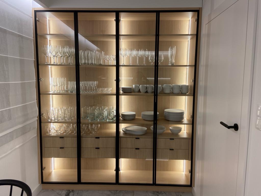 Wysoka, przeszklona szafa z drewnianymi szufladami i wewnętrznym oświetleniem, wypełniona szkłem i porcelaną, z czarnymi ramami i uchwytami, stojąca obok białych drzwi.