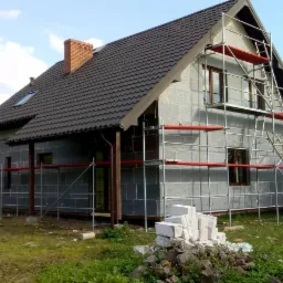 Dom jednorodzinny w trakcie prac związanych z ociepleniem ścian zewnętrznych, widoczne szare płyty izolacyjne i rusztowanie z czerwonymi elementami zabezpieczającymi, na pierwszym planie stos...
