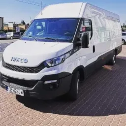 Biały bus marki Iveco na kostce brukowej, widok z przodu i boku, tablica rejestracyjna CB 428JW, w tle budynki i drzewa.