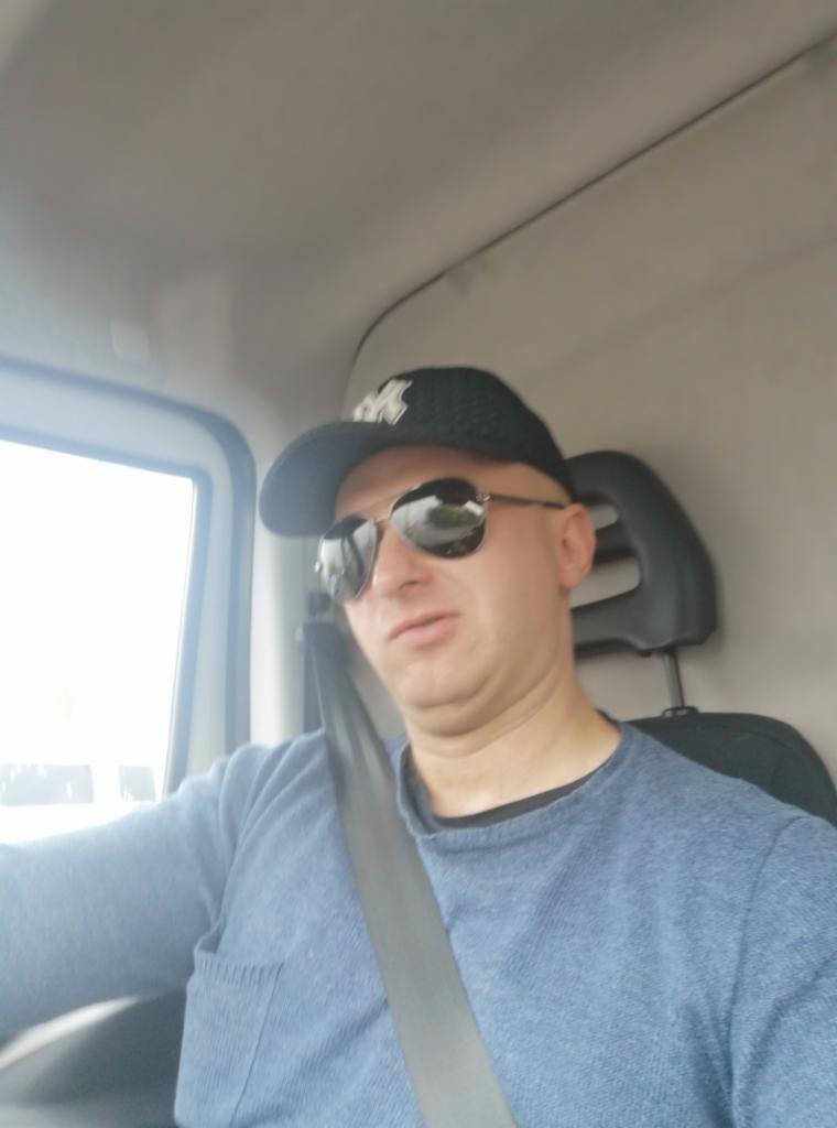 Kierowca w czapce z daszkiem i okularach przeciwsłonecznych, zapięty pasem bezpieczeństwa, siedzący w kabinie busa z widocznym oknem.