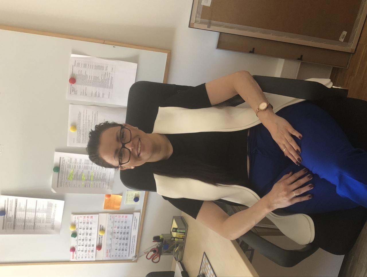 Uśmiechnięta kobieta w okularach siedzi na krześle biurowym w gabinecie, w tle tablica z dokumentami i kalendarz.