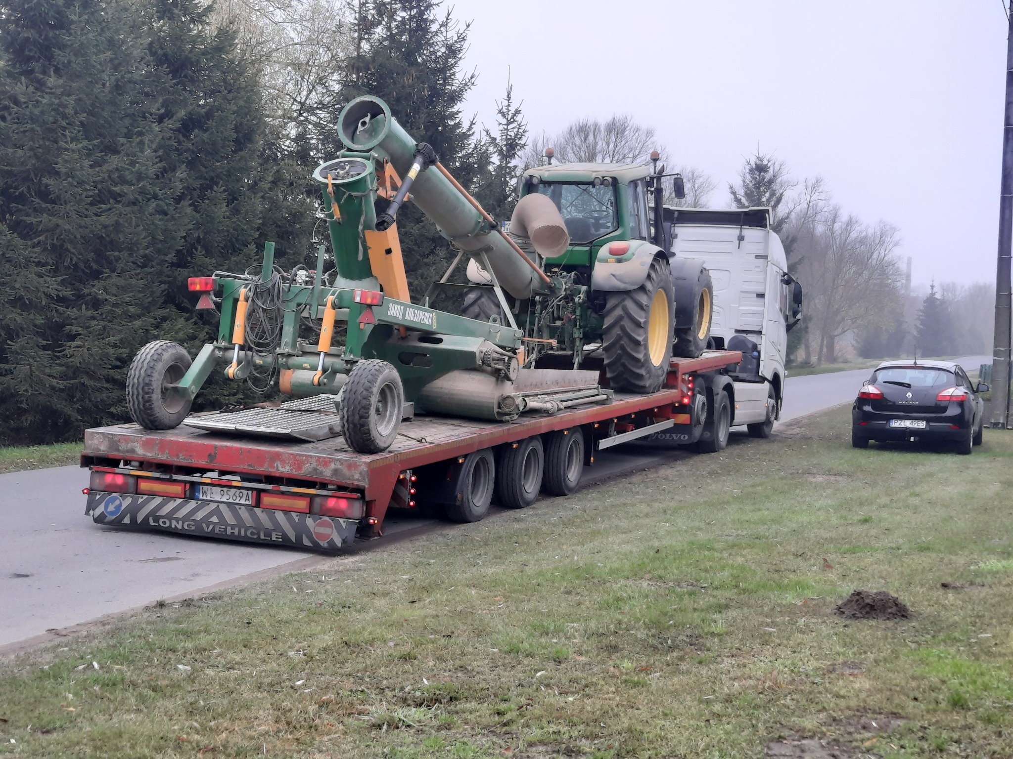 Zielony traktor i maszyna rolnicza marki ZAWOD KOBZARENKA przewożone na niskopodwoziowej ciężarówce Volvo, widok z boku na drodze w pochmurny dzień.
