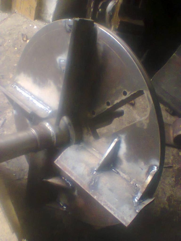 Wieloelementowa, spawana konstrukcja stalowa z okrągłą tarczą, widoczne spawy i ślady obróbki, w tle warsztatowy nieporządek.