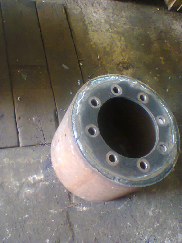 Metalowy cylinder z kołnierzem i otworami na śruby, leżący na zabrudzonej podłodze warsztatu, widoczne ślady obróbki.