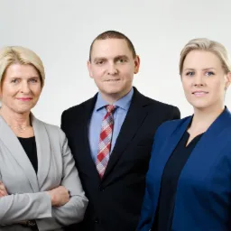 Portret grupowy: kobieta w szarym żakiecie ze skrzyżowanymi rękami, mężczyzna w czarnym garniturze i krawacie w kratę, kobieta w niebieskim żakiecie na jasnym tle.