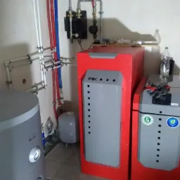 Kotłownia z dwoma piecami PIK, zbiornikiem na wodę i instalacją hydrauliczną z rurami w kolorach czerwonym i niebieskim, widoczne elementy armatury i osprzętu.