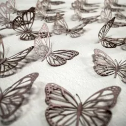Delikatne, wycinane laserowo motyle 3D w odcieniach różu i szarości, rozmieszczone na teksturowanej, białej powierzchni, tworzące dekoracyjny wzór.