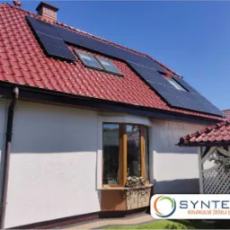 Dom z czerwonym dachem, na którym zamontowano panele słoneczne, widoczne okna dachowe, błękitne niebo w tle.