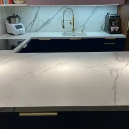 Nowoczesna kuchnia z blatem imitującym marmur, złotą armaturą i granatowymi szafkami, ekspres do kawy i robot kuchenny na blacie.