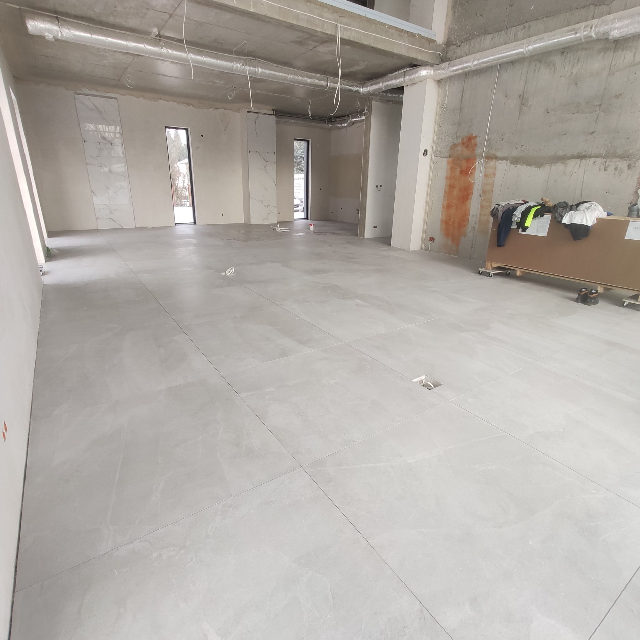 Jasnoszara, matowa podłoga z płytek w surowym wnętrzu, widoczne pionowe fragmenty płytek na ścianach i instalacje pod sufitem.