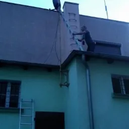 Mężczyzna na drabinie ustawia antenę satelitarną i telewizyjną na dachu budynku z zieloną elewacją, widoczne kable i maszt antenowy.