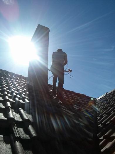 Monter anten stoi na dachu z dachówki ceramicznej, obok komina, pod silnym słońcem, instalując antenę satelitarną.