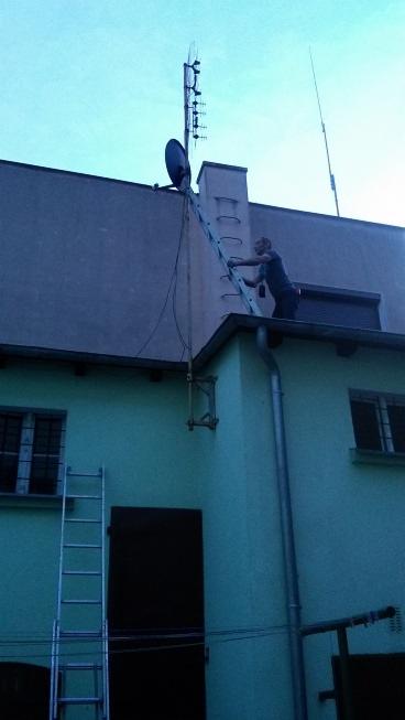 Mężczyzna na drabinie ustawia antenę satelitarną i telewizyjną na dachu budynku z zieloną elewacją, widoczne kable i maszt antenowy.