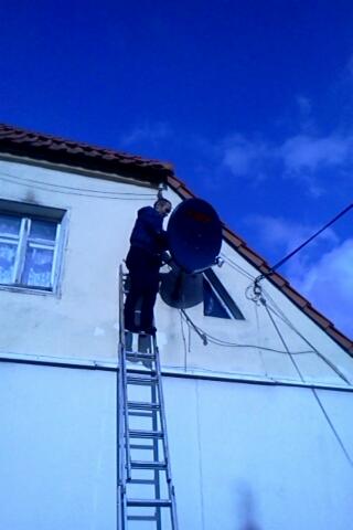 Instalator na drabinie montuje antenę satelitarną na elewacji domu z oknem, widoczne kable i niebieskie niebo.