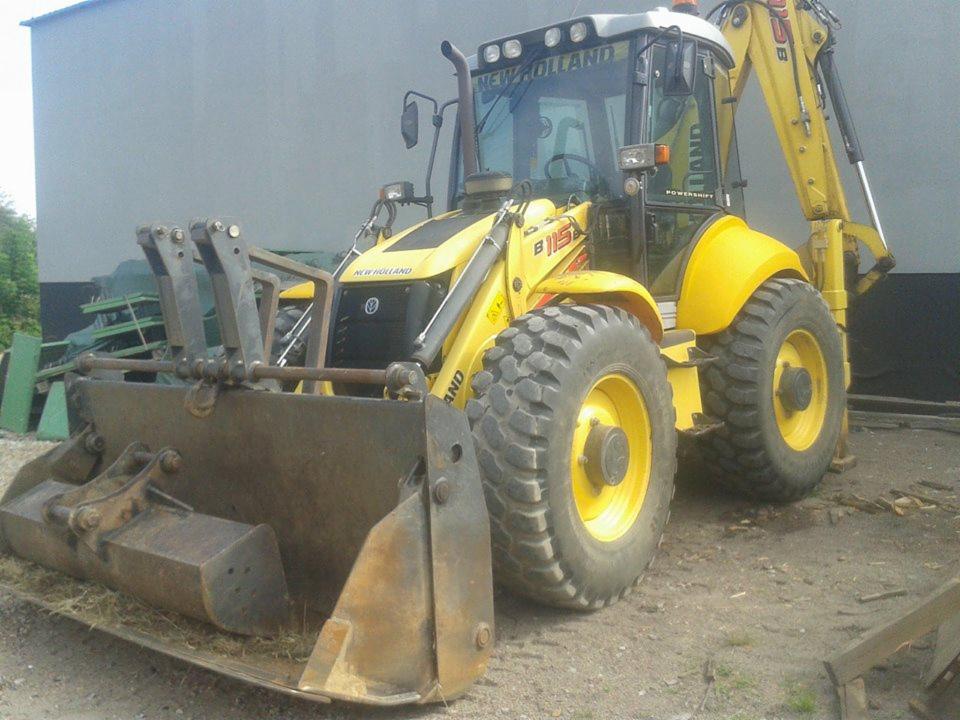 Żółta koparko-ładowarka New Holland B115 z łyżką i widłami na tle szarego budynku i zieleni. Maszyna stoi na ziemi, gotowa do pracy.