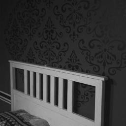 Kamil Spólnik - Ściana w sypialni z ciemnym, wzorzystym zdobieniem w stylu damask, widoczna za białym zagłówkiem łóżka z pościelą w geometryczne wzory. Dwie poziome linie w kolorze zagłówka biegną wzdłuż dolnej...