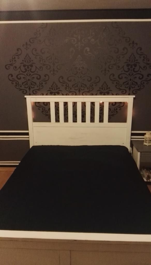 Ciemna ściana w sypialni z ornamentowym wzorem w odcieniach szarości, biała rama łóżka z czarną narzutą, nocna lampka na stoliku.