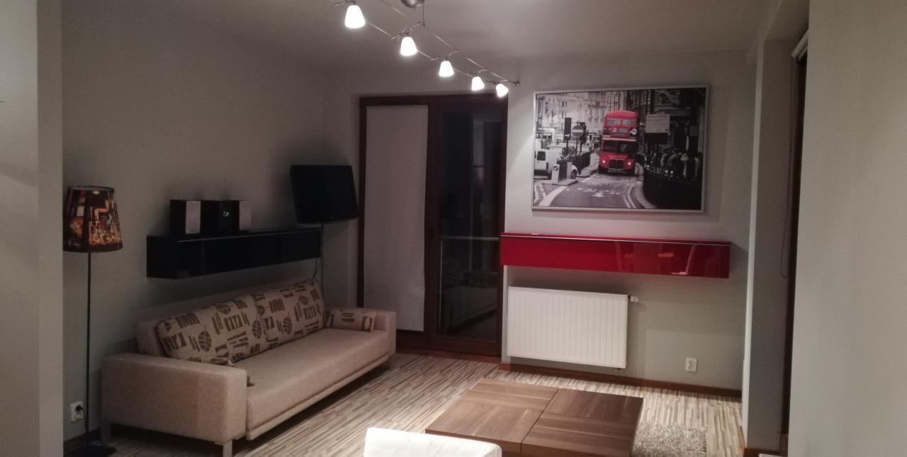 Jasne wnętrze salonu po odświeżeniu, z szarą ścianą, na której wisi obraz z motywem Londynu, czerwona szafka i biały grzejnik.