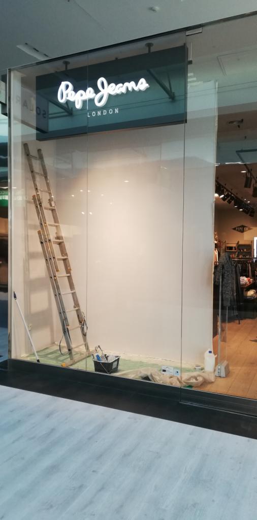 Malowanie wnętrza sklepu Pepe Jeans London: drabina oparta o świeżo pomalowaną białą ścianę, widoczny pojemnik z narzędziami malarskimi i folia ochronna na podłodze.