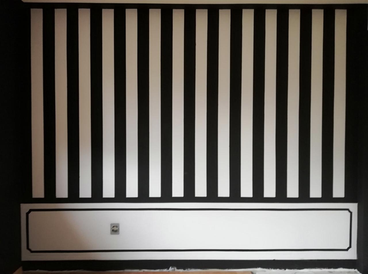 Ściana w czarno-białe pionowe pasy, z białą lamperią obramowaną czarną linią i gniazdkiem elektrycznym