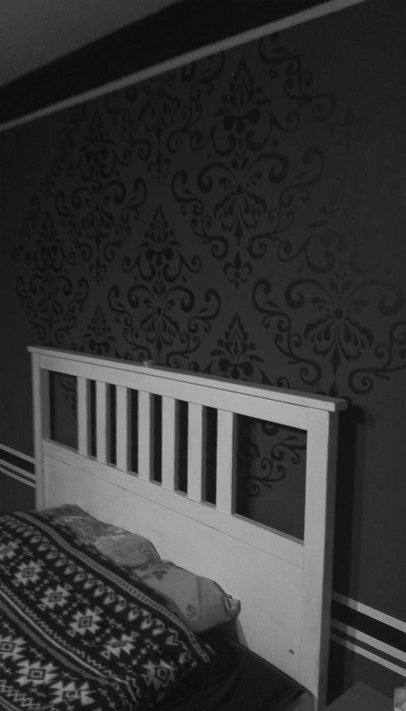 Ściana w sypialni z ciemnym, wzorzystym zdobieniem w stylu damask, widoczna za białym zagłówkiem łóżka z pościelą w geometryczne wzory. Dwie poziome linie w kolorze zagłówka biegną wzdłuż dolnej...