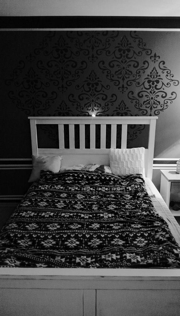 Czarno-białe zdjęcie sypialni z wzorzystą ścianą za białym łóżkiem i kołdrą w etniczne wzory. Ściana ozdobiona dużym, ciemnym ornamentem roślinnym, z górną i dolną krawędzią podkreśloną poziomymi...
