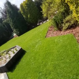 Ułożony trawnik z rolki na zboczu, z obrzeżami z kory i roślinami w donicach z betonu.