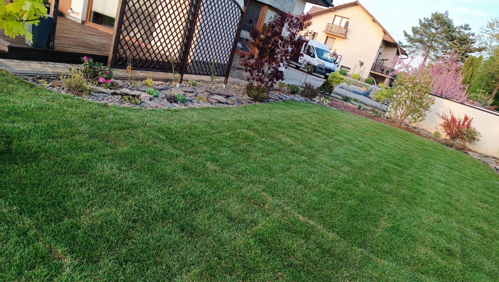 Perfekcyjnie utrzymany trawnik z niską trawą, rabaty z kamieniami i roślinami ozdobnymi, w tle dom z drewnianym tarasem i zaparkowany biały samochód dostawczy.