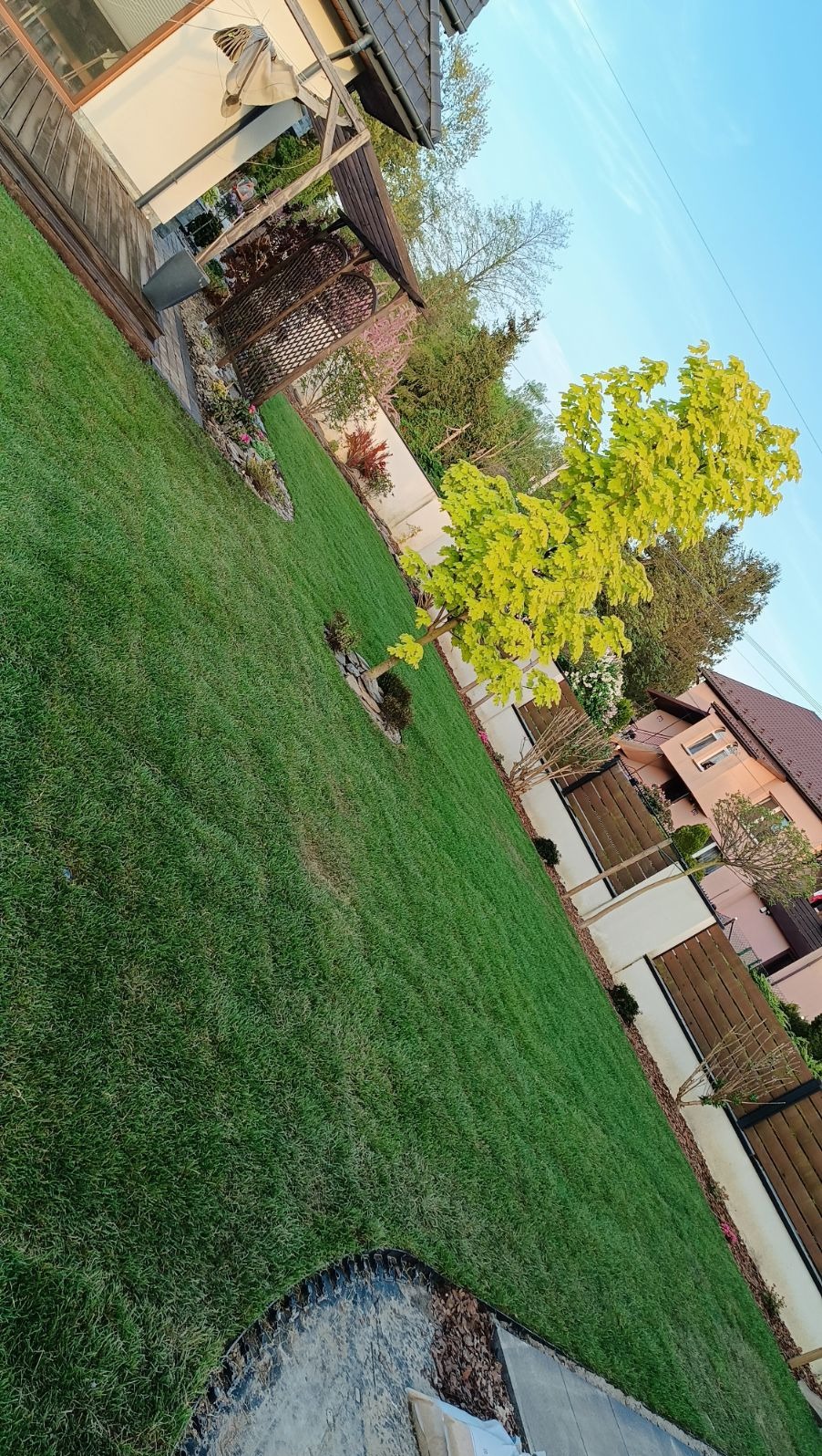 Starannie utrzymany trawnik z młodymi drzewami i rabatami kwiatowymi, widok na dom z brązowym dachem i drewnianym ogrodzeniem, pergola w tle.