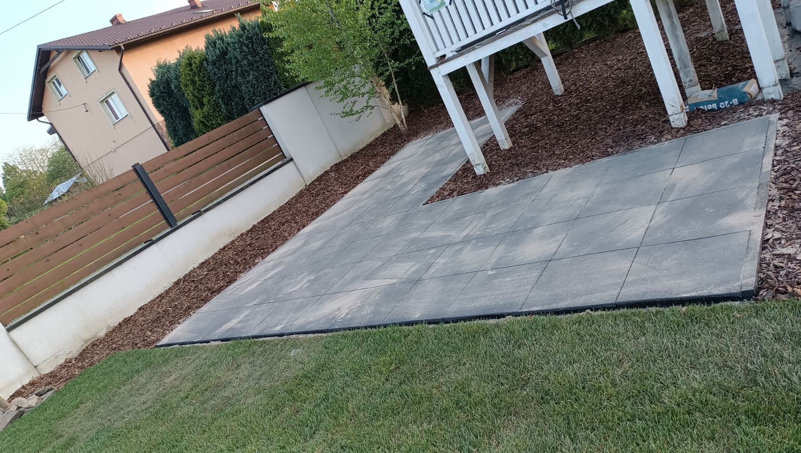 Ułożona szara kostka brukowa na podwórku z elementami drewnianej architektury ogrodowej, obrzeża z kory, trawa i ogrodzenie w tle.