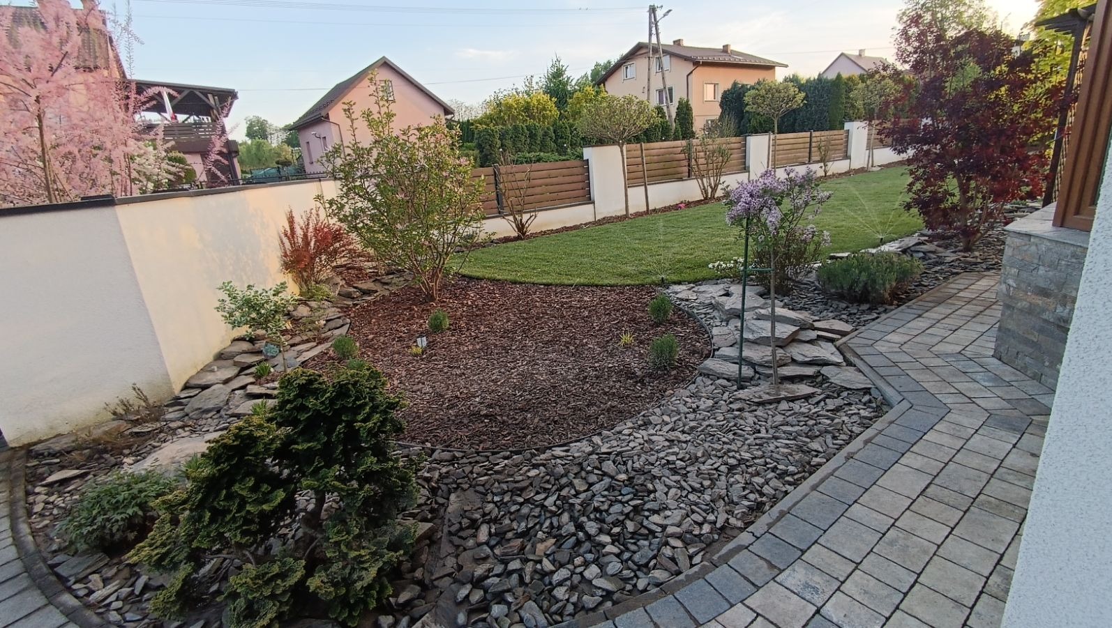 Ujęcie z poziomu chodnika na zadbany ogród z trawnikiem, rabatami wysypanymi korą i kamieniami, oraz systemem nawadniania włączonym w tle. Ogród otoczony murem i drewnianym płotem.