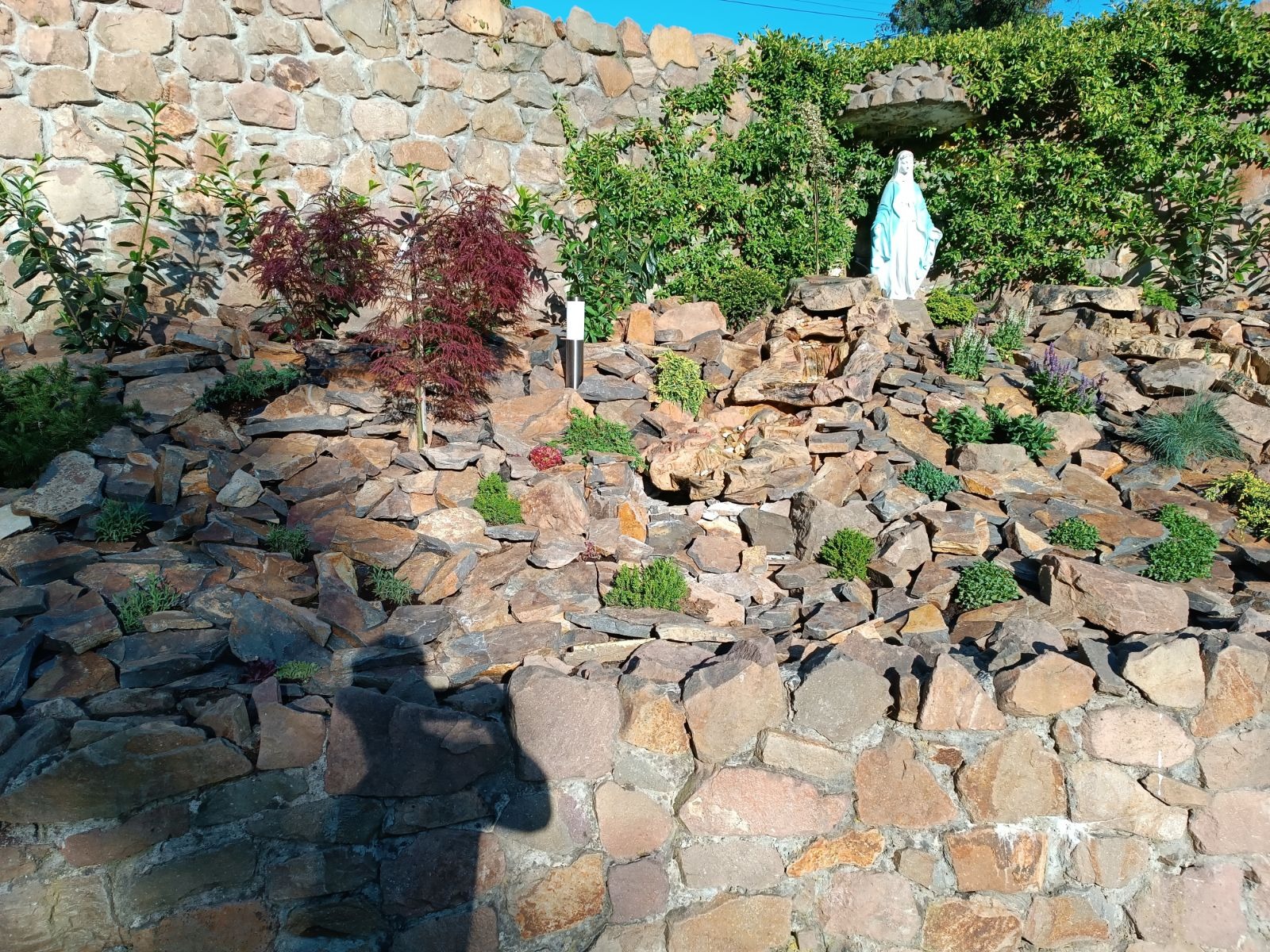 Ogród skalny z kaskadą wodną, roślinami ozdobnymi i figurką Matki Boskiej, otoczony kamiennym murem.