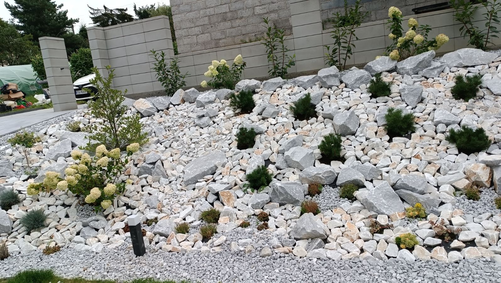 Nowoczesny ogród skalny z jasnymi kamieniami, karłowatymi iglakami i kwitnącymi hortensjami na zboczu, z widocznym fragmentem betonowego muru i zielonym trawnikiem w tle.