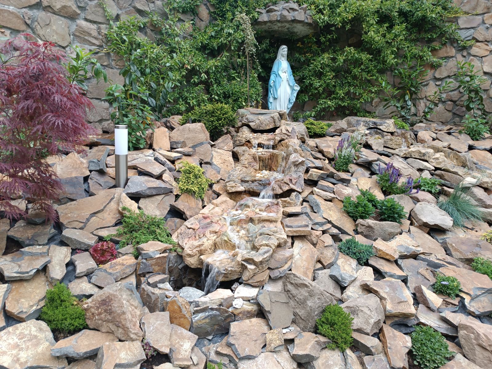 Ogród skalny z kaskadowym strumieniem, figurą Matki Boskiej w tle i ozdobnym oświetleniem, zróżnicowana roślinność.