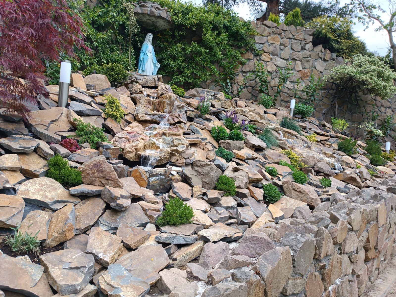 Ogród skalny z kaskadą wodną i figurą Matki Boskiej, oświetlony lampami, z różnorodną roślinnością.